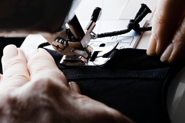 Sağdan sola, siyah bir ev yapımı yüz örtüsüne ya da yüz maskesine dikiş atılır. Yaşlı yıpranmış ellerin rehberliğinde.