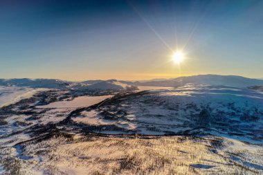 Norveç 'in karlı kış dağları üzerinde, huş ağaçları ve çam ağaçları ile günbatımı altın ışık, sakin mavi gökyüzü. Atoklinten dağ bölgesi, Joesjo, Lappland belediyesi, Kuzey İsveç