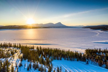 Norveç 'in karlı kış dağları ve Rossvatnet Gölü, çam ve huş ağaçları üzerinde gün batımı altın ışık, sakin mavi gökyüzü. Hattfjelldal Belediyesi, Kuzey Norveç