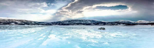 Panorama of frozen lake with small island in Northern Mountains starting to melt and water flows on the ice surface. Blue skies and sun rays light down. Joesjo lake in Lappland, Northern Sweden.