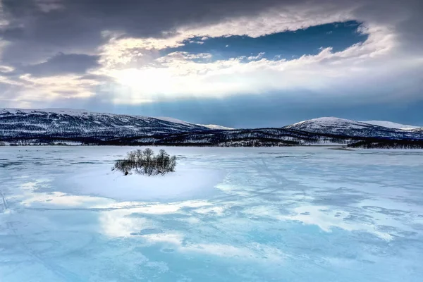 Frozen lake with small island in Northern Mountains starting to melt and water flows on the ice surface. Blue skies and sun rays light down. Joesjo lake in Lappland, Northern Sweden.