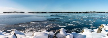 Baltık Sis 'teki kış sahili manzarası, açık deniz, yüzen buz, kıyı şeridindeki büyük taşları kaplayan taze kar, mavi gökyüzü, güneşli gün, hafif esinti, çam ağacı ormanlı adalar. Kuzey İsveç, Umea