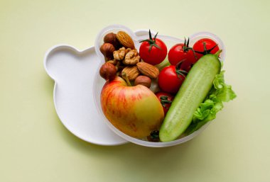 Children's lunch box shaped bear head with vegetarian food: vegetables, fruits, nuts. With space for text, on a light yellow background. Concept of healthy food, snack for children clipart