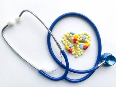 Mavi steteskop bükülmüş halkalar, içinde kalp, renkli haplarla kaplanmış: ağrı kesiciler, vitaminler, balık yağı, beyaz desenli kağıt arka planda gastrik madde.