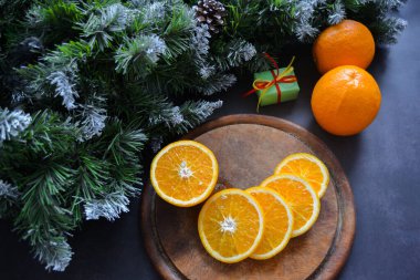 Eski bir ahşap kesim tahtasında dilimlenmiş sulu portakallar, iki tam meyve, yanında hediye olan küçük bir kutu. Sol üstte don, kar ve koni ile kabarık ladin dalları var. Noel ruhu