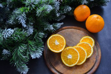 Ağaç kesme tahtasının üzerinde iki meyve yan yana dilimlenmiş sulu olgun portakallar. Sol üstte don, kar ve koni ile kabarık ladin dalları var. Noel konsepti, yeni yıl.