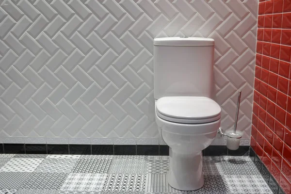 Beyaz ve kırmızı seramik duvarlı kusursuz temiz banyo, grafik desenli siyah ve beyaz zemin, drenaj tanklı beyaz klozet ve yanında fırça.