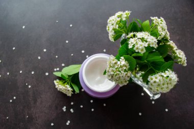 Beyaz çiçekli ve yeşil yapraklı Spirea dalları altında beyaz kremalı, narin bir leylak kavanozu var. Beyaz çiçek yaprakları koyu bir zemine saçılmış.