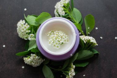 Beyaz çiçeklerin içinde beyaz desenli beyaz kremalı leylak kavanozu. Etrafta çiçekli ve yeşil yapraklı Spirea dalları var. Koyu arkaplan