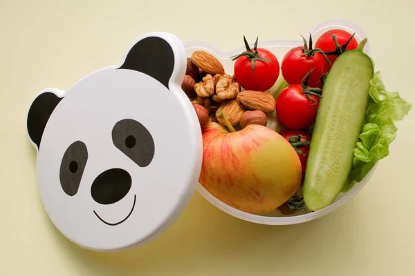 Children\'s lunch box shaped panda head with vegetarian food: vegetables, fruits, nuts. With space for text, on a light yellow background. Concept of healthy food, snack for children