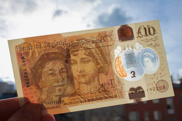 El, polimer plastikten yapılmış yepyeni bir 10 pound tutuyor. Kraliçe ve Jane Austen 'in portresi ve banknotun mavi arka planında, şeffaf bir pencereden yansıyan güneş ışığı.