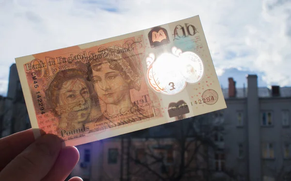 El, polimer plastikten yapılmış yepyeni bir 10 pound tutuyor. Kraliçe ve Jane Austen 'in portresi ve banknotun mavi arka planında, şeffaf bir pencereden yansıyan güneş ışığı.
