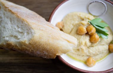 Maydanozla süslenmiş humus ve seramik kasede nohut ve ahşap arka planda bir parça ekmek.