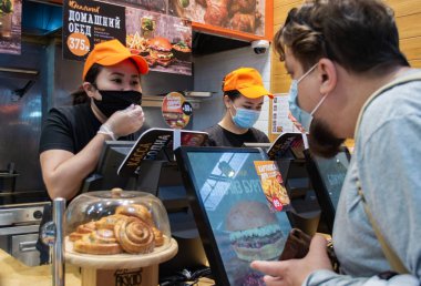 Moskova, Rusya, Temmuz 2020: Sakallı ve tıbbi maskeli bir adam Burger Cafe 'den sipariş veriyor. Siyah koruyucu maskeli ve eldivenli kafe çalışanlarının arkasında.