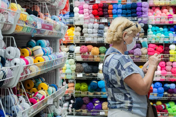 stock image Moscow, Russia, August 2020: a Woman in a protective medical mask in a craft store among rows of colorful threads and yarns made of cotton, wool