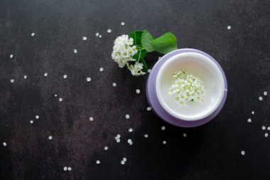 Beyaz çiçeklerin içinde beyaz desenli beyaz kremalı leylak kavanozu. Etrafta çiçekli ve yeşil yapraklı Spirea dalları var. Karanlık arka plan. Boşluğu kopyala