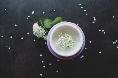 Beyaz çiçeklerin içinde beyaz desenli beyaz kremalı leylak kavanozu. Etrafta çiçekli ve yeşil yapraklı Spirea dalları var. Koyu arkaplan.