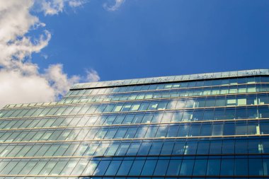 Modern bir ofis binasının köşesinde güneş parlıyor. Bulutlu mavi gökyüzü aynı pencerelerin camına yansıyor..