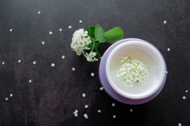 Beyaz çiçeklerin içinde beyaz desenli beyaz kremalı leylak kavanozu. Etrafta çiçekli ve yeşil yapraklı Spirea dalları var. Koyu arkaplan.