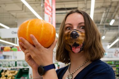 Moskova, Rusya, Ağustos 2020: Köpek suratlı komik maskeli genç bir kadın, markette parlak turuncu bir balkabağı tutuyor. Virüsten korunma ve Cadılar Bayramı için hazır olma kavramı..