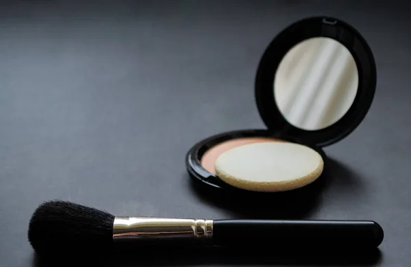 A black makeup brush next to an open box with a mirror and powder puff and pink powder. Black background. Copy space.