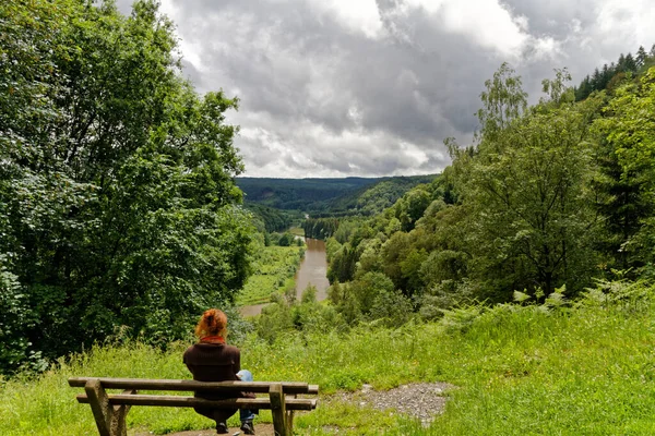 Young caucasian woman on a bench on the top of a mountain overlooking river valley with green trees and river in Belgium