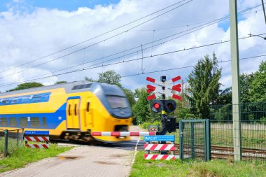 Hollanda sarısı yüksek hızlı şehirlerarası treni kapalı bariyerler ve yanıp sönen kırmızı ışıklarla kapalı bir demiryolu geçidini geçiyor. Mavi uyarı işareti metni.
