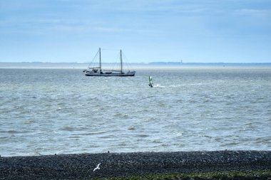 Harlingen, Hollanda, 23 Temmuz 2020: Rüzgâr sörfçüsü ve eski geleneksel yelkenli gemisi, siyah taş hendek, rıhtım ve Ufukta Terschelling adası karakteristik deniz feneriyle