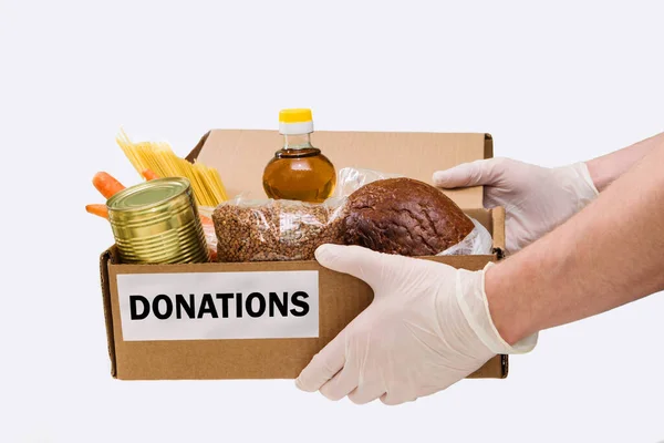 Las manos de los hombres en guantes médicos dan una caja de cartón con comida, donación de alimentos, entrega a domicilio, horizontal, espacio para copiar. Trigo sarraceno, pan, pasta, conservas, mantequilla, arroz, cebollas, zanahorias . — Foto de Stock