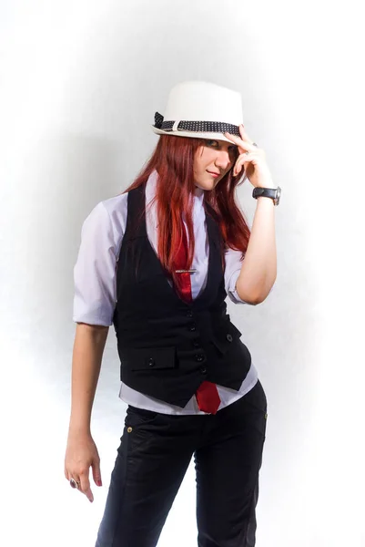 Una hermosa joven pelirroja con una blusa blanca, chaleco negro y corbata roja y un sombrero blanco sobre un fondo blanco, con la cabeza hacia abajo, mira desde debajo del borde de su sombrero . — Foto de Stock