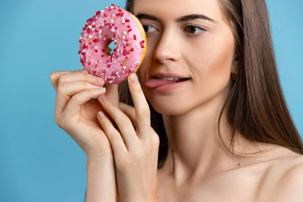 Крупним планом жінка з довгим волоссям тримає пончик і стирчить язиком. Жіноча модель пончик перед обличчям на синьому фоні . — стокове фото