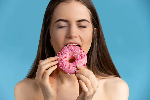 Дівчина з закритими очима їсть рожеві пончики. Смачний пончик на ізольованому синьому фоні — стокове фото