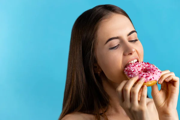 Крупним планом портрет приємної красивої дівчини, що їсть рожеві пончики, ізольовані на синьому фоні — стокове фото