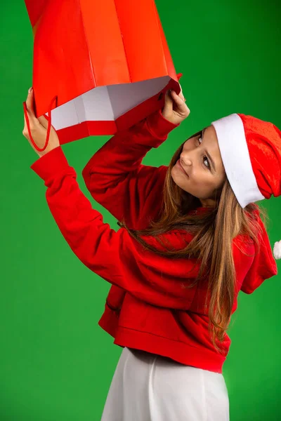 Красива молода дівчина в капелюсі Санта і Різдвяний светр дивиться в торговельну сумку на ізольованому зеленому тлі — стокове фото