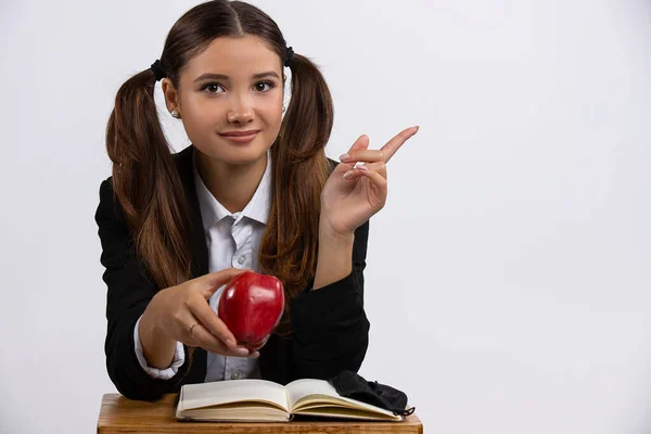 Školní koncept. Portrét usměvavé školačky sedící u stolu s ukazováčkem nahoru u jablka, bílé pozadí — Stock fotografie