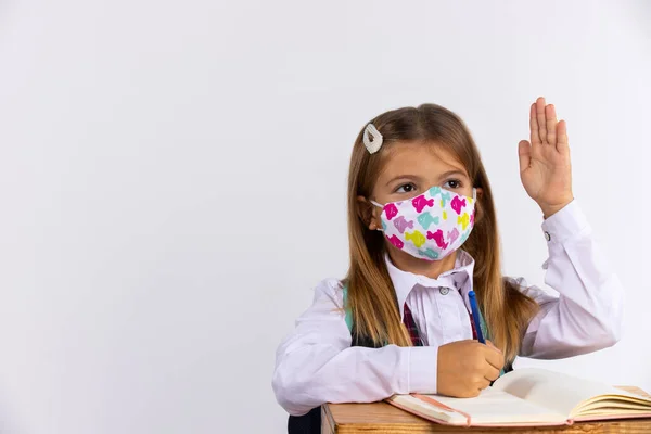 Colegiala con máscara médica, levantó las manos en la lección, entrenando libros en la mesa. Concepto escolar, copiar espacio vacío. — Foto de Stock