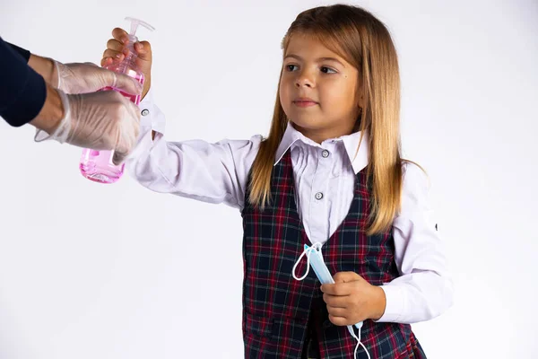 Niña confundida con máscara medicinal en la mano y uniforme escolar tratar de tomar el desinfectante en la mano — Foto de Stock