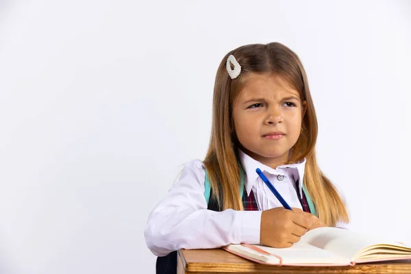 穿着校服、穿着紧身衣的小女孩孤零零地坐在书桌前，白色背景。女学生不喜欢做作业. — 图库照片