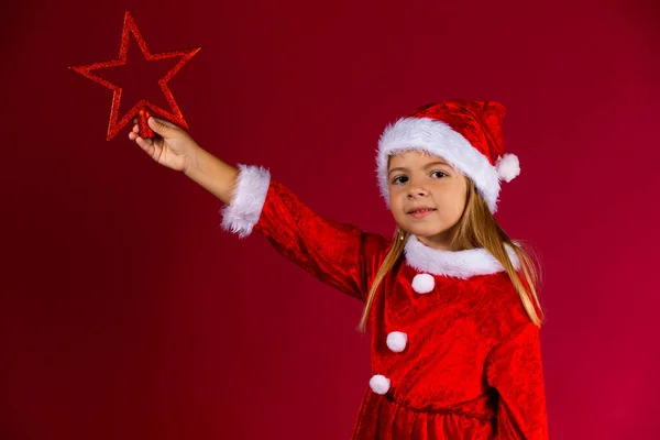 Hermosa niña en Santa vestido y sombrero sostiene la estrella roja de Navidad, aislado sobre fondo rojo — Foto de Stock