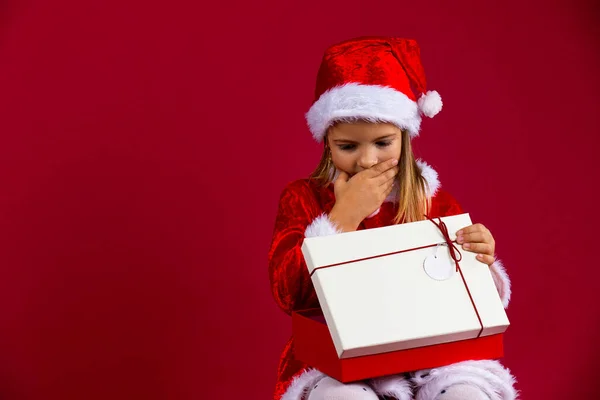 Kırmızı arka planda izole edilmiş, Noel Baba şapkası ve kostüm giymiş, açık bir hediye kutusu olan düşünceli genç bir kızın portresi. — Stok fotoğraf