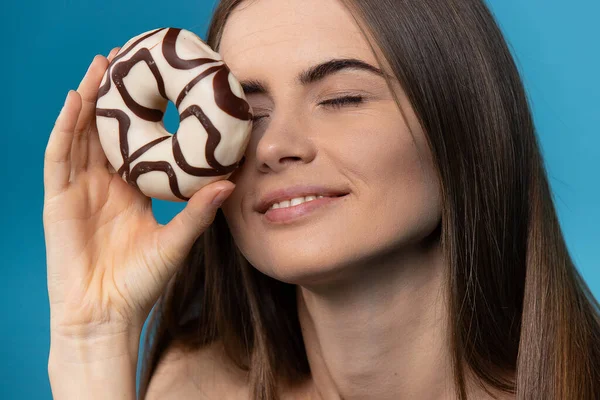 Спокуса їжі. Досить біла жінка з закритими очима насолоджується шоколадним пончиком на ізольованому на синьому фоні . — стокове фото