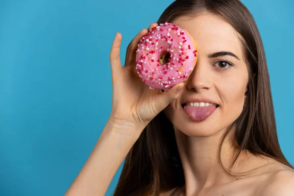 Портрет красивої дівчини з рожевим глазурованим пончиком, дивиться на камеру з язиком. Студійний знімок синього ізольованого фону . — стокове фото