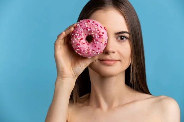 Досить кавказька модель дівчина приховує своє око з рожевими глазурованими пончиками. Спокуса їжі. Ізольовані на синьому фоні — стокове фото