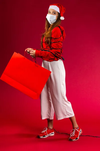 Mujer joven con suéter rojo y paquete en sus manos, con una máscara está cubierta con guirnalda mirando a la cámara — Foto de Stock