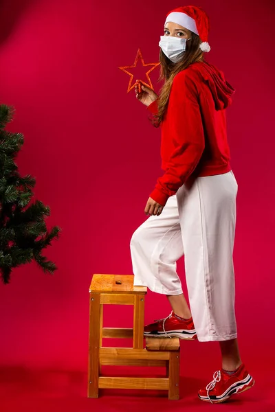 Mujer caucásica con máscara médica, sube a la silla para decorar el árbol de Navidad con una estrella roja. Fondo rojo — Foto de Stock