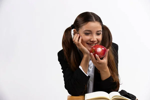 Vzrušená studentka sedící u lekce ve svém dřevěném stole a usmívající se na své červené jablko. Izolované bílé pozadí — Stock fotografie