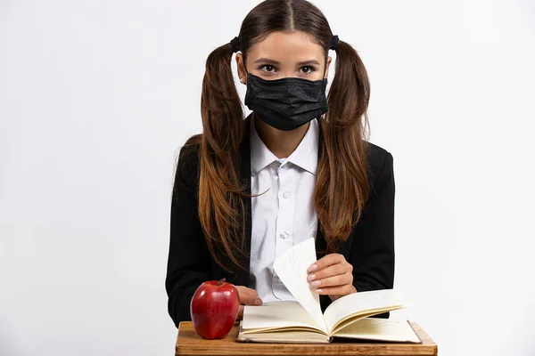 Studioaufnahme einer jungen Frau mit schwarzer Schutzmaske, die am Schreibtisch sitzt und durch das Buch blättert. Weißer Hintergrund — Stockfoto