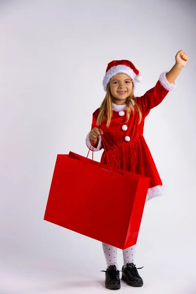 Girl with gift bag on Christmas shopping. Smiling kid in winter clothes holding red shopping bags. White background