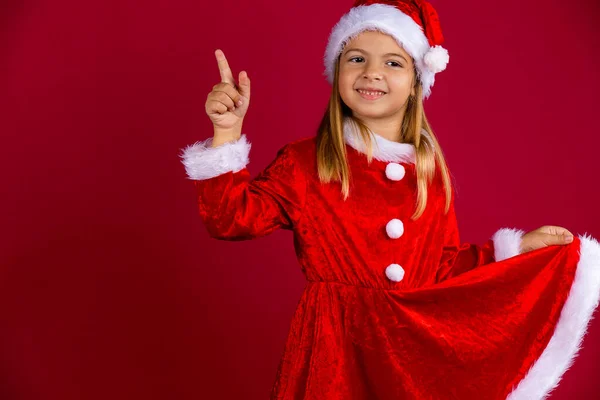 Retrato de chica linda en traje de Santa Claus y sombrero de invierno. Niño sosteniendo el vestido rojo a mano y señalando hacia arriba. — Foto de Stock