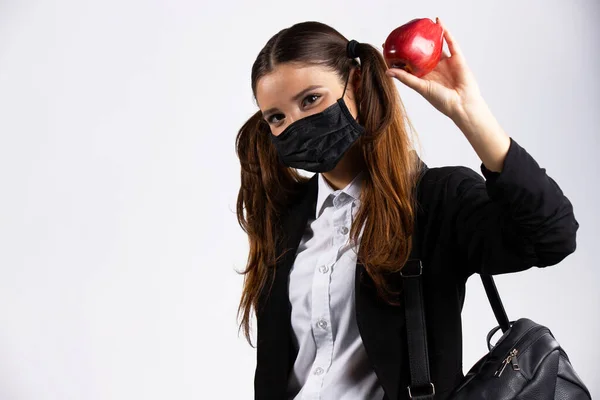 Kaukasisches Mädchen mit Maske im Gesicht hält einen Apfel in der Hand und eine Tasche auf der Schulter blickt in die Kamera — Stockfoto